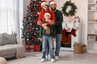 Mutlu ebeveynler ve Noel Baba şapkalı küçük oğulları Noel arifesinde evde sarılıyorlar.