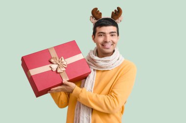 Geyik boynuzları takmış mutlu genç adam yeşil arka planda Noel hediyesiyle