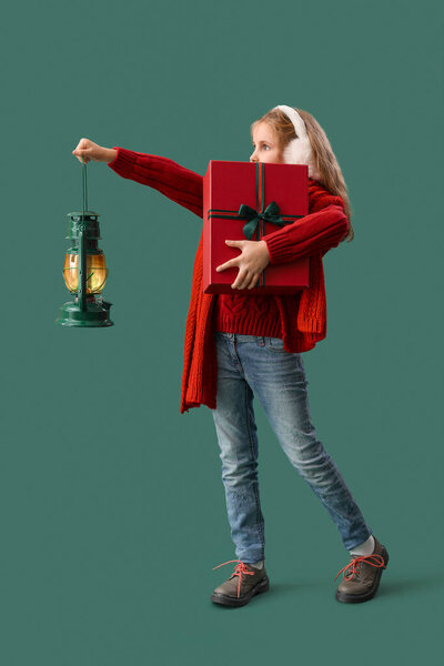 Little girl in earmuffs with Christmas gift and lantern on green background