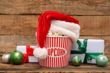 Noel Baba şapkalı bir kova patlamış mısır, Noel topları ve tahta arka planda hediyeler.