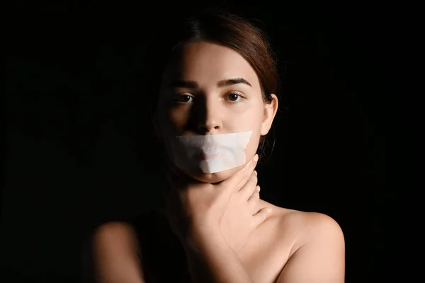 Young woman with taped mouth on dark background. Censorship concept