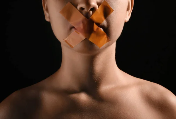 Young woman with taped mouth on dark background, closeup. Censorship concept