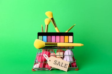 Makyaj fırçaları, süngerler, göz falı ve yeşil arka planda satış etiketi olan alışveriş sepeti.