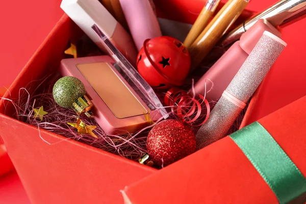 Christmas gift box with makeup products, balls and bell on red background, closeup