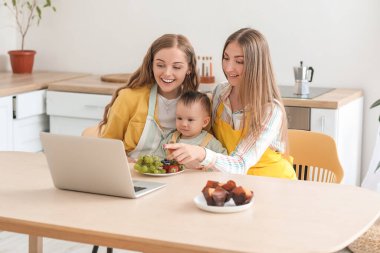 Küçük bebekleriyle mutfaktaki masada çizgi film izleyen mutlu lezbiyen çift.