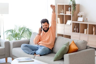 Evdeki gri koltukta cep telefonuyla konuşan mutlu genç sakallı adam.
