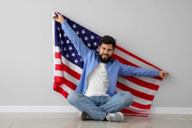Amerikan bayrağı taşıyan yakışıklı sakallı adam gri duvarın yanında oturuyor.