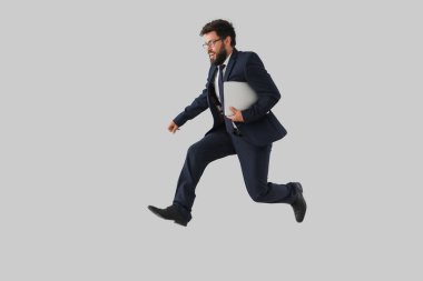 Gri duvarın yanında laptopuyla atlayan yakışıklı sakallı iş adamı.