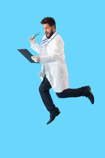 带着剪贴板和眼镜跳上蓝色背景的令人惊讶的男医生 — 图库照片