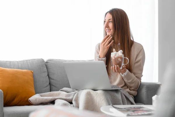 Young woman with glass cup of cocoa and laptop sitting on sofa at home