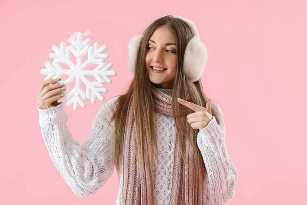 穿着温暖的耳罩的年轻女子指着粉色背景的大雪花 — 图库照片