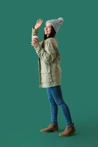 若いです女性で暖かいです帽子とともにコーヒーカップ手を振って緑の背景 — ストック写真