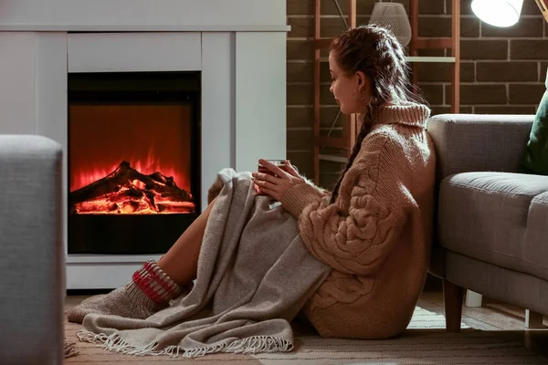 Woman with cup of tea sitting near fireplace at home