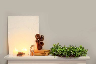 Mumlarla dolu boş bir poster, Noel çelengi, çam kozalakları ve gri duvarın yanındaki şömine üzerindeki kitaplar.