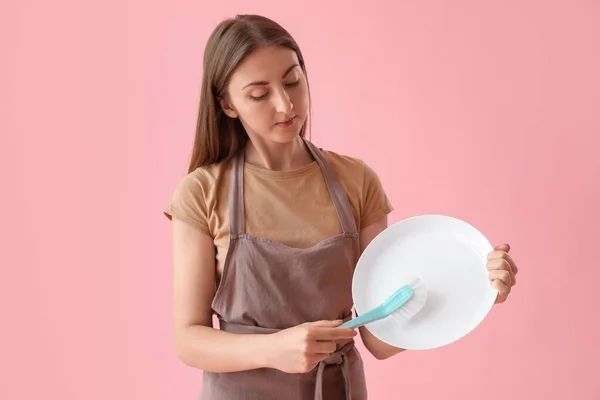Young woman cleaning plate with brush on pink background