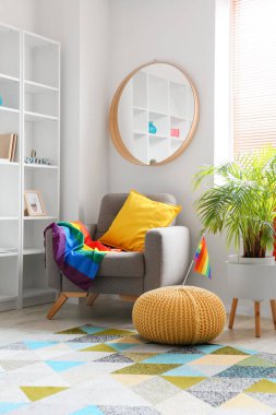 Beyaz duvarın yanında ayna, çiçek ve gökkuşağı bayraklı rahat bir koltuk.