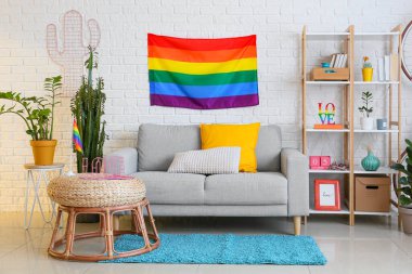 Rahat kanepe, raf ünitesi, ev bitkileri ve beyaz tuğla duvarın yanında gökkuşağı bayrakları.