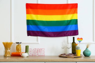 Masada gözlüklü bir şişe şarap ve beyaz duvarda asılı gökkuşağı bayrağı