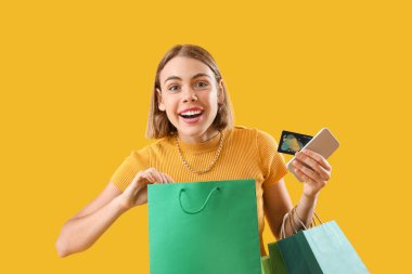 Sarı arka planda kredi kartı, cep telefonu ve alışveriş torbaları olan mutlu genç bir kadın.