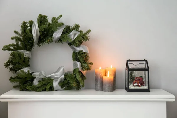 Christmas wreath with burning candles and gift on mantelpiece near light wall