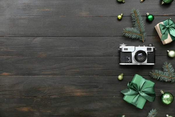 Photo camera with Christmas branches, balls and gifts on black wooden background