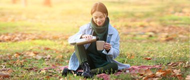 Genç bir kadın sonbahar parkında termostan fincana çay dolduruyor.