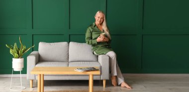 Yeşil duvarın yanındaki kanepede oturmuş çay içen olgun bir kadın.