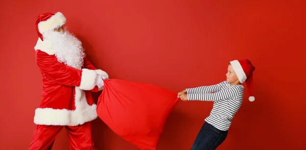 Niegrzeczny Chłopiec Próbuje Wziąć Torbę Prezenty Świętego Mikołaja Czerwonym Tle — Zdjęcie stockowe