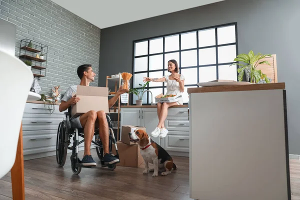 ピザを持つ若い女性 車椅子の夫と移動中の日にキッチンで犬 — ストック写真