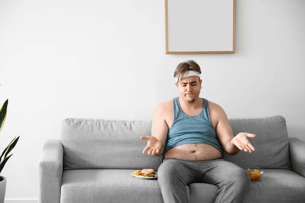 若いです太りすぎ男とともに不健康な食べ物座っていますソファに家 — ストック写真