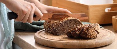 Woman cutting fresh bread on table in kitchen, closeup