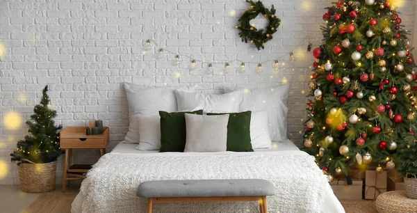 Interior of bedroom with Christmas trees, wreath and glowing lights hanging on light wall