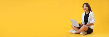 Sarı arka planda dizüstü bilgisayarı olan genç Asyalı bir kadın.