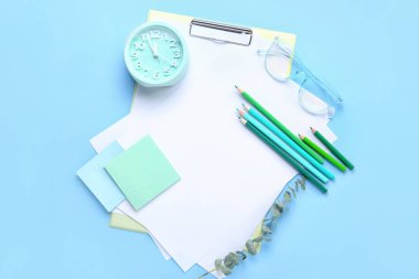 Boş kağıt kağıtlar, kalemler, çalar saat, yapışkan notlar ve mavi arkaplanda gözlük olan pano.