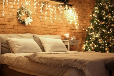 Karanlık yatak odasının içinde Noel çelengi, köknar ağacı ve parlayan ışıklar var.