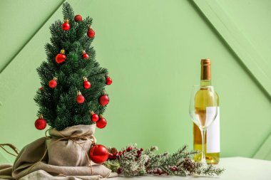 Yeşil duvarın yanındaki masada cam ve şarap şişesiyle küçük bir Noel ağacı.