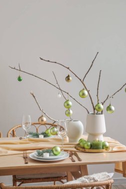 Ağaç dalları olan vazolar ve gri duvarın yanındaki yemek masasında yeşil Noel topları.