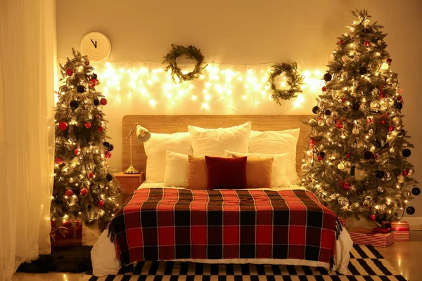 Interior of bedroom with Christmas trees, wreaths and glowing lights at night