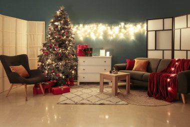Karanlık oturma odasının içinde Noel ağacı, çekmeceler ve parlayan ışıklar var.