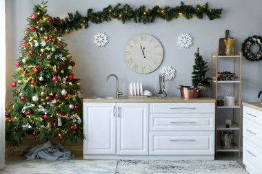 Mutfağın içinde Noel ağaçları, beyaz tezgahlar ve büyük bir saat var.