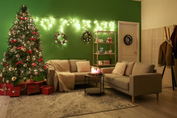 Interior of dark living room with glowing Christmas tree, wreaths and sofas