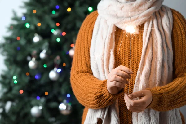 Woman in scarf with Christmas sparkler against blurred fir tree, closeup