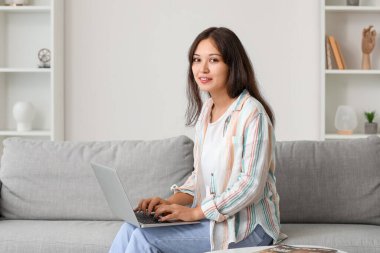 Genç Asyalı kadın evdeki kanepede dizüstü bilgisayar kullanıyor.