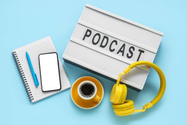 Kelime PODCAST, kulaklıklar, bir fincan kahve, cep telefonu ve mavi arka planda not defteri olan pano