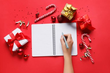 Boş defter, kalem, Noel hediyeleri ve kırmızı arka planda süslemeleri olan kadın eli.