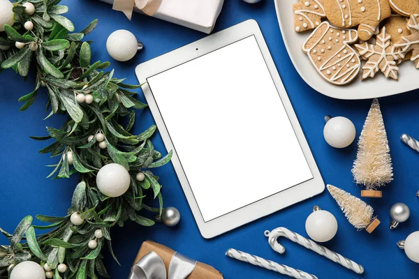 Blank tablet computer with Christmas mistletoe wreath, cookies and decor on blue background