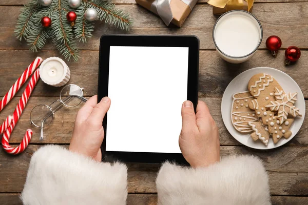 Santa Claus with tablet computer, glass of milk, cookies and Christmas decor on wooden background