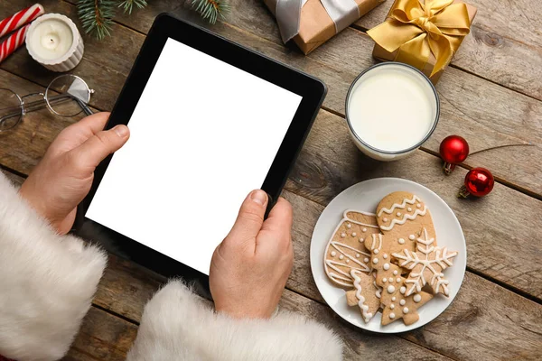 Santa Claus with tablet computer, glass of milk, cookies and Christmas decor on wooden background