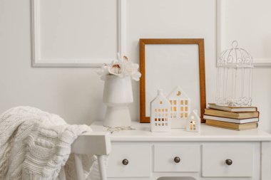 Ev şeklinde mum tutucular, çiçekli vazo, çerçeve ve kitaplar ışık duvarının yanındaki masada.