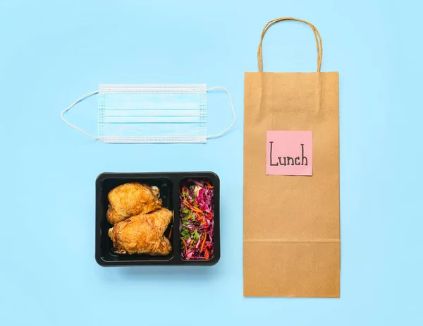Food box with delicious meal, medical mask, paper bag and sticky note with word LUNCH on color background